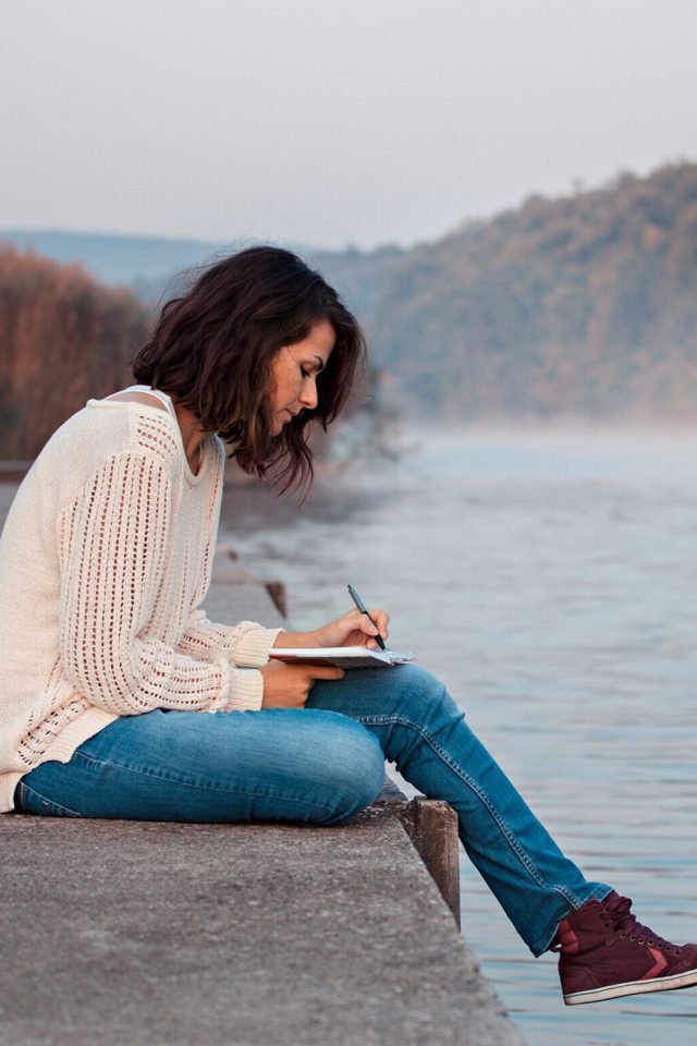 Girl writing diary by the lake in the early cold morning.
