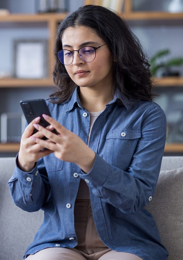 Young Indian woman in shirt and glasses sitting on sofa at home and using mobile phone.