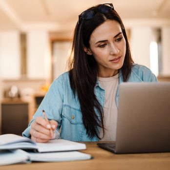 Focused charming woman writing down notes while working with laptop at home