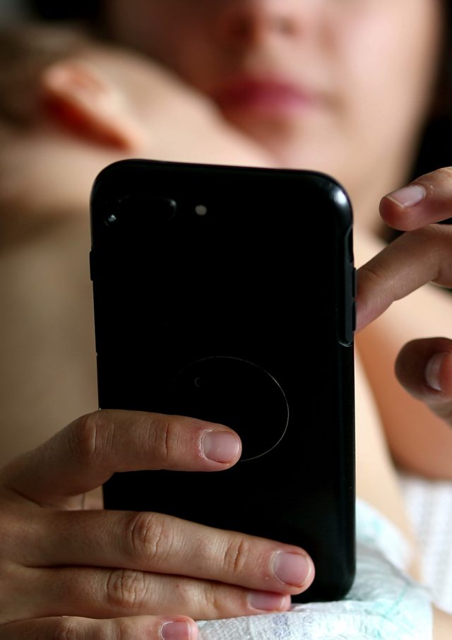 Casual mother using cellphone device in bedroom with sleeping baby infant napping